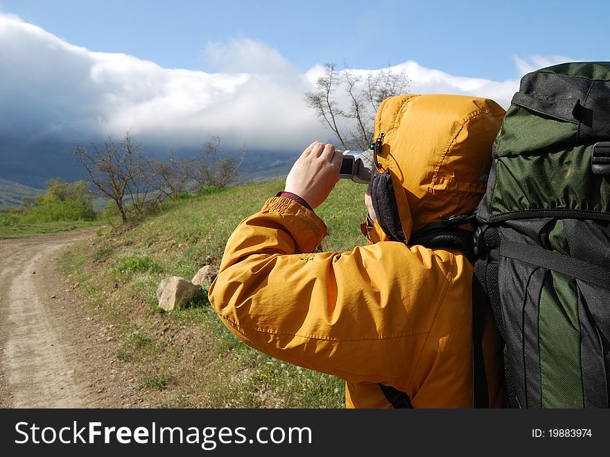 Hiker is shooting a landscape by video camera. He is standing on the natural road. He is carrying a large rucksack. Hiker is shooting a landscape by video camera. He is standing on the natural road. He is carrying a large rucksack.