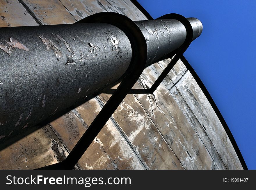 Large iron pipe strapped to side of community water tank with blue sky above. Large iron pipe strapped to side of community water tank with blue sky above