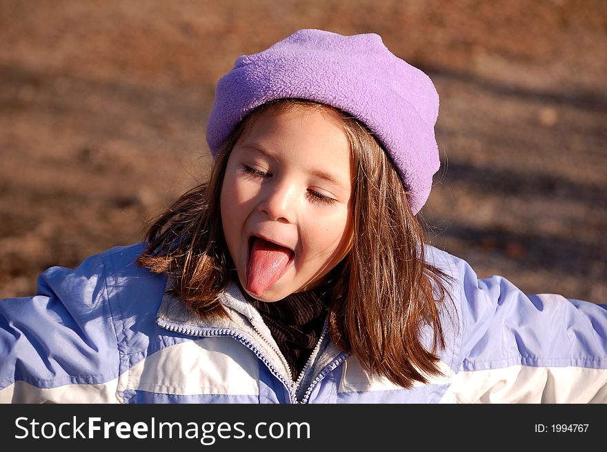 Four year old girl dressed in a purple hat and coat. She is sticking out her tongue with her arms stretched out and the late autumn sun is on her face. Four year old girl dressed in a purple hat and coat. She is sticking out her tongue with her arms stretched out and the late autumn sun is on her face.