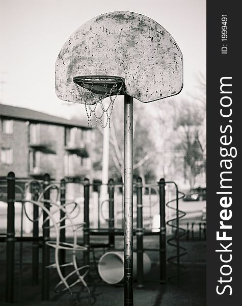 School-yard basketball hoop at sunset with playground blurred behind. Black and white.