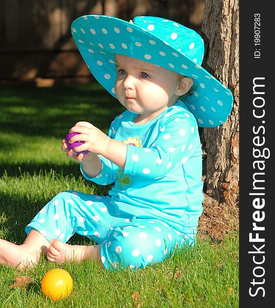 A toddler sits on the grass while wearing her light blue polkadot hat and swimsuit playing with purple and yellow balls. A toddler sits on the grass while wearing her light blue polkadot hat and swimsuit playing with purple and yellow balls