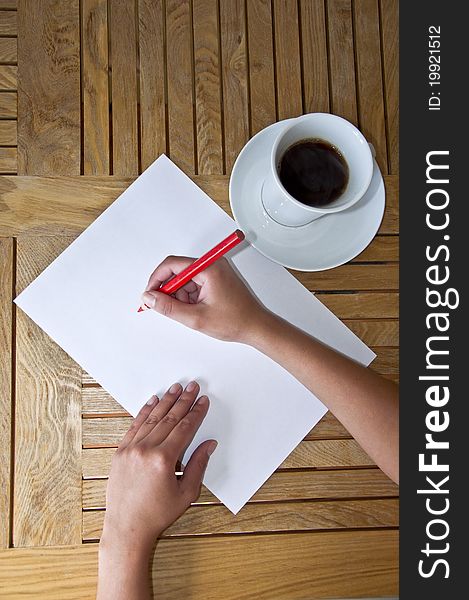 Female hand with pencil writing on a white sheet of paper. Wooden table and a cup of black coffee