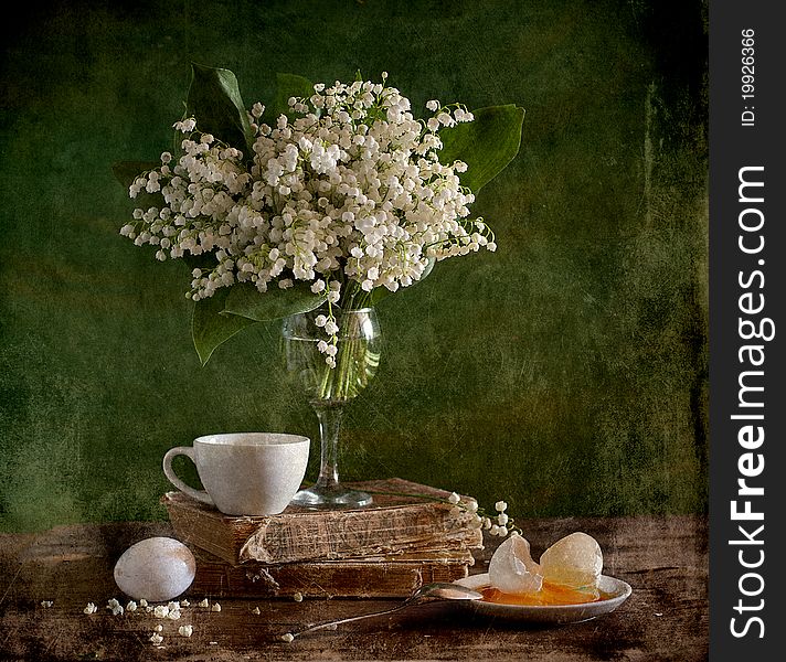 A bouquet of lilies of the valley is in glass, alongside book, broken egg and cup
