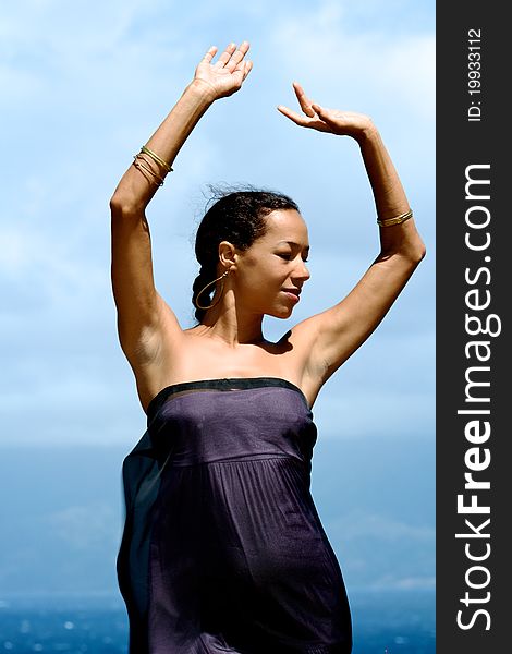 An exotic Hawaiian woman in a flowing dress has arms upraised to the sun. She is standing on a cliff overlooking the ocean. Vertical image orientation. An exotic Hawaiian woman in a flowing dress has arms upraised to the sun. She is standing on a cliff overlooking the ocean. Vertical image orientation.
