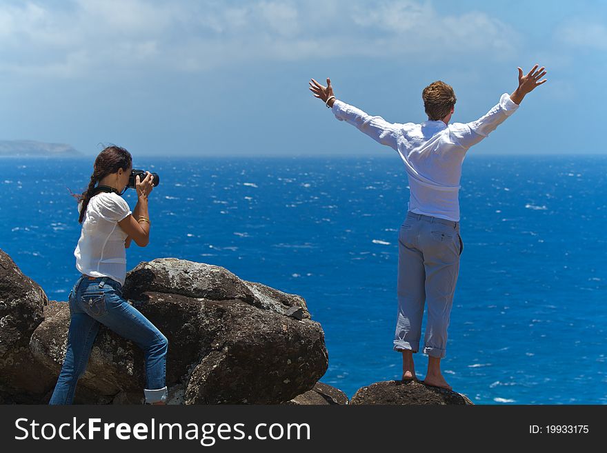 With outstretched arms, a man stands on a rock at the top of a cliff overlooking the ocean. A woman is photographing him from behind. With outstretched arms, a man stands on a rock at the top of a cliff overlooking the ocean. A woman is photographing him from behind.