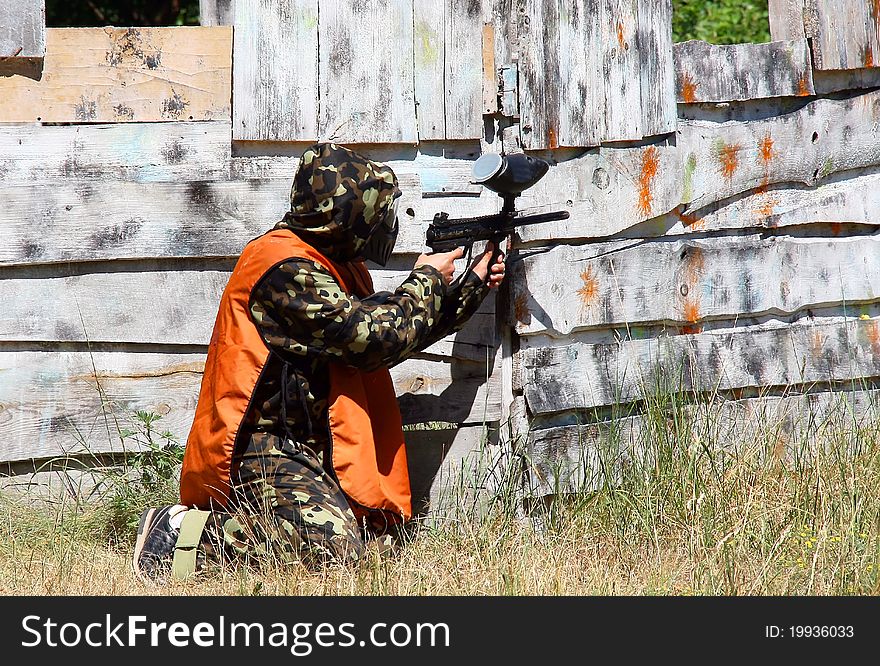 The man in camouflage and a mask shoots from the marker at a wooden wall. The man in camouflage and a mask shoots from the marker at a wooden wall