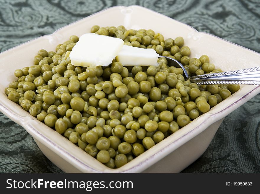 Cooked peas with butter in a ceramic dish on a green background with spoon.
