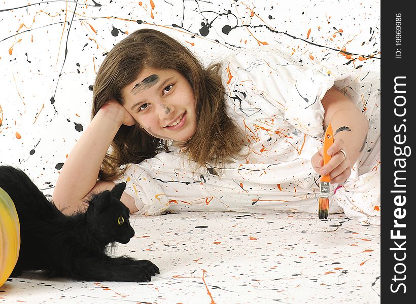 An attractive preteen painting in an orange and black paint spattered smock against a paint-spattered background. A black cat and pumpkins nearby. An attractive preteen painting in an orange and black paint spattered smock against a paint-spattered background. A black cat and pumpkins nearby.