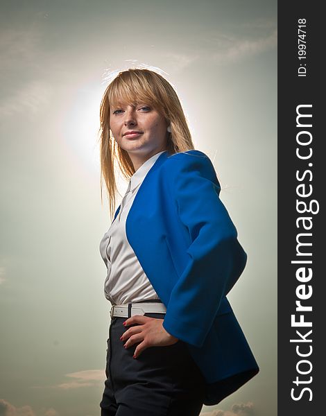 Blond in white shirt and blue jacket looking into future on cloudy sky background. Sun lighting her hair. Blond in white shirt and blue jacket looking into future on cloudy sky background. Sun lighting her hair