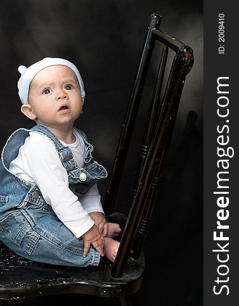 Image of adorable baby wearing denim overalls sitting on a black chair. Image of adorable baby wearing denim overalls sitting on a black chair