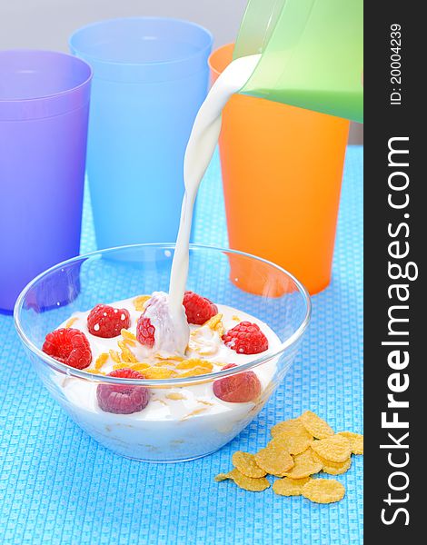 Cornflakes in glass bowl with berries, milk,  and colorful plastic cups on a blue background