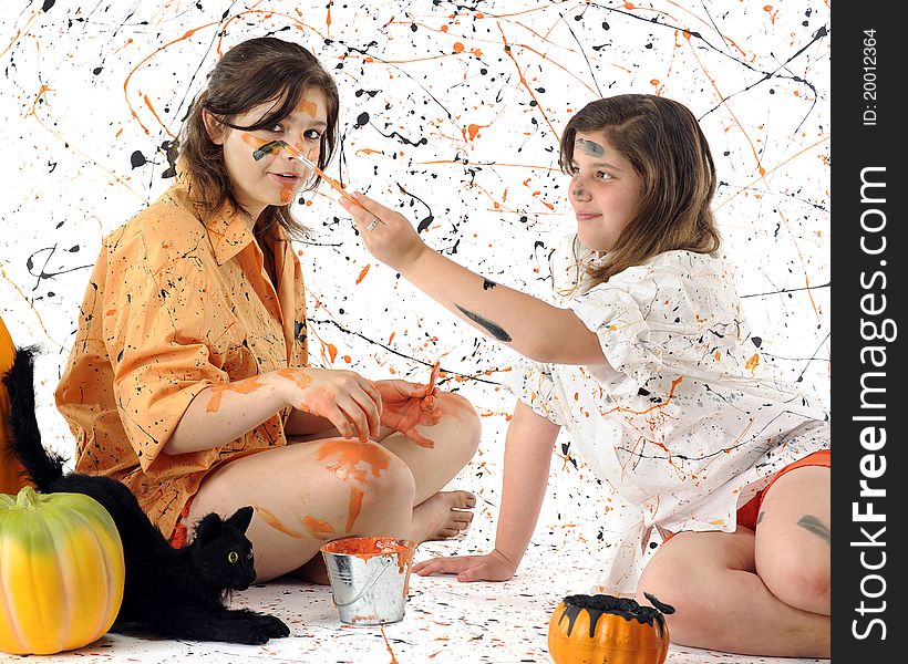 A young teen and her tween sister making a mess of themselves with black and orange paint. Pumpkins and a black cat nearby. Some motion blur on the brush painting the girl's face. A young teen and her tween sister making a mess of themselves with black and orange paint. Pumpkins and a black cat nearby. Some motion blur on the brush painting the girl's face.