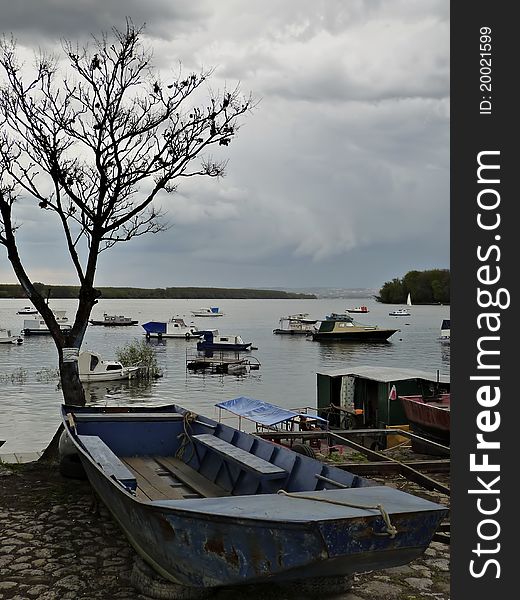 Old fishing boat at river bank, leafless tree in background, cloudy sky before storm.