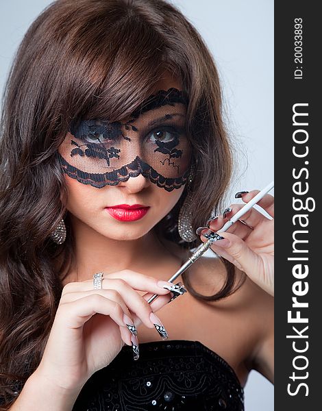 Closeup portrait of attractive young brunette girl with long dark ringlets and fine art manicure wearing lacy mask on her eyes holding graceful silver mouthpiece with thin ladies' cigaret on blue background. Closeup portrait of attractive young brunette girl with long dark ringlets and fine art manicure wearing lacy mask on her eyes holding graceful silver mouthpiece with thin ladies' cigaret on blue background