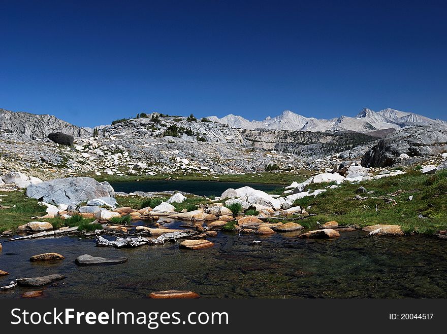 Two clear alpine lakes in the High Sierra with rugged white peaks in the background, contrasted against a dark blue sky.