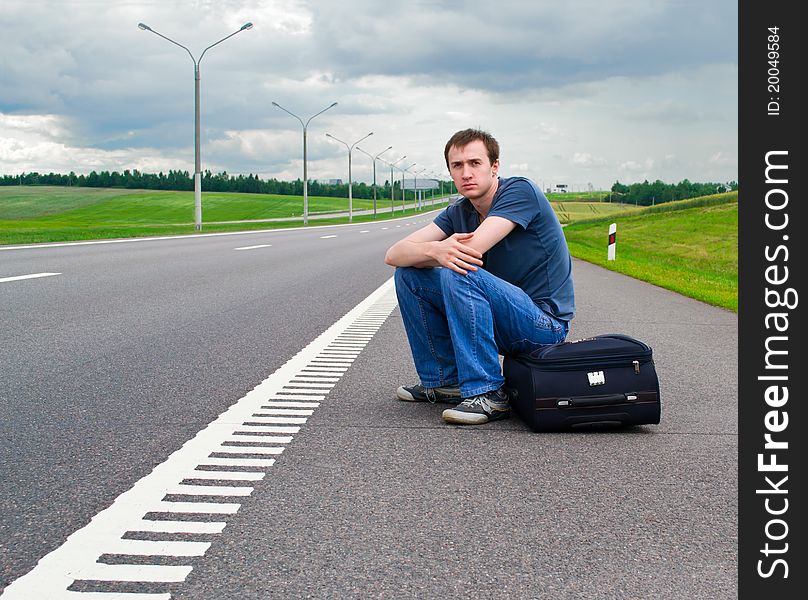 The young man sits pending on road with a suitcase