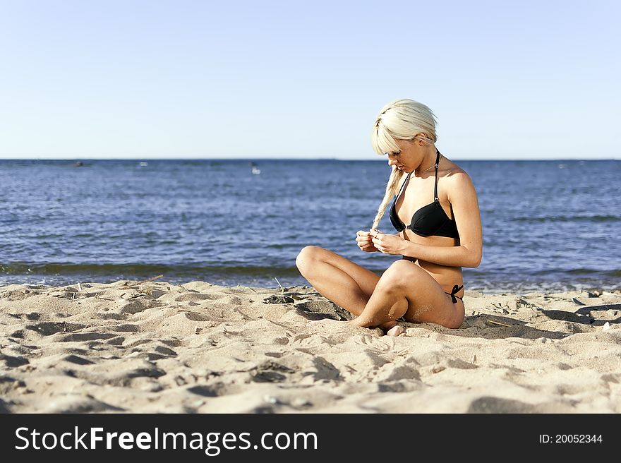 Blonde girl in a black bathing suit sitting on shore blue sea on the sand. Blonde girl in a black bathing suit sitting on shore blue sea on the sand