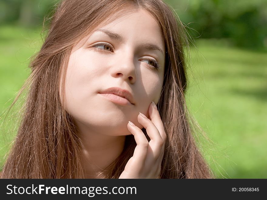 Portrait of young woman with clear skin is touching her face by hand in park on green background. Portrait of young woman with clear skin is touching her face by hand in park on green background