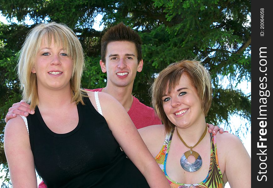 Three friends, young man with hands on shoulders of 2 young women outside with spruce tree in the backgroungd. Head and shoulders. Three friends, young man with hands on shoulders of 2 young women outside with spruce tree in the backgroungd. Head and shoulders.