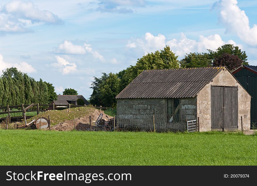 An old barn with wooden doors in a Dutch landscape. Green grass in the foreground. The sky is blue with white clouds. Some green trees in the background.