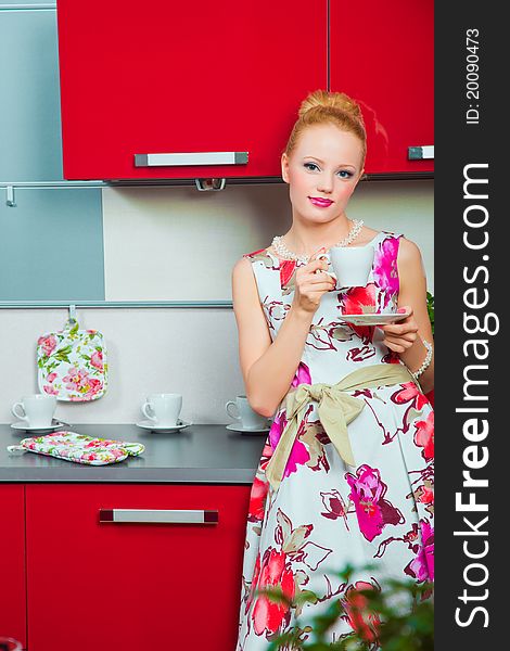 Young beautiful woman wearing white and pink dress with cup of morning coffee in interior of red modern kitchen. Young beautiful woman wearing white and pink dress with cup of morning coffee in interior of red modern kitchen