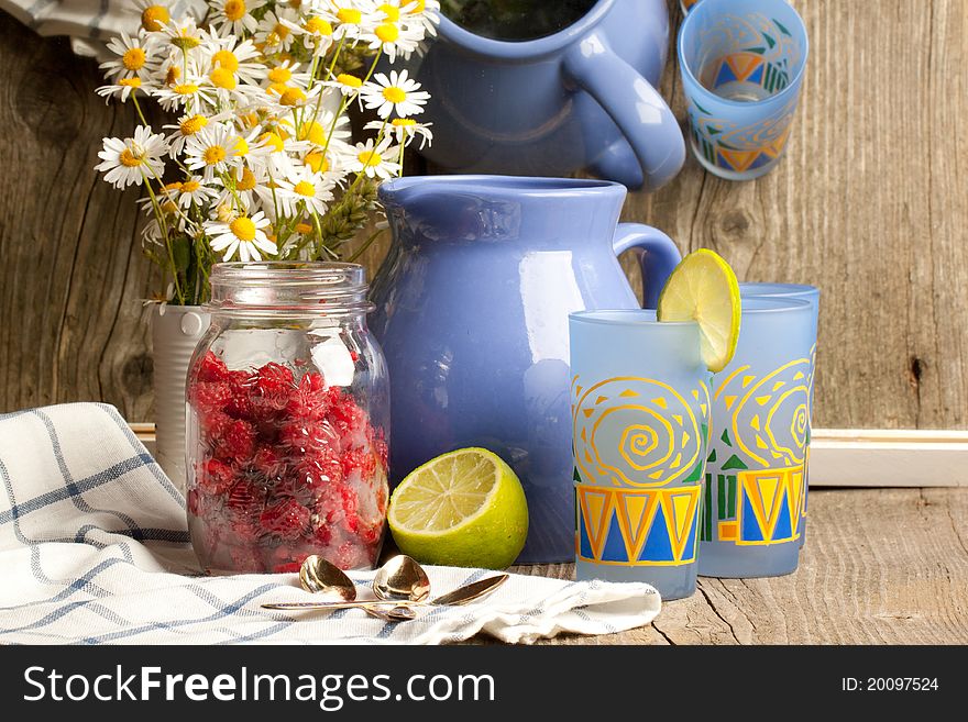 Composition with jug and glasses of lemonade, fresh ripe raspberries, half of lime and bunch of camomile on old wooden table near mirror with reflex. Composition with jug and glasses of lemonade, fresh ripe raspberries, half of lime and bunch of camomile on old wooden table near mirror with reflex