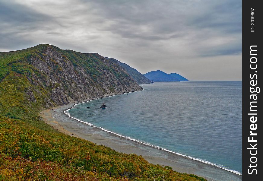 The dark clouds on whole sky above the capes and sea. Dusk. Autumn. The yellow bushes and grey beach with line of white surf are on foreground. Japanese sea, Russian Far East, Primorye, Tasovaya bay. The dark clouds on whole sky above the capes and sea. Dusk. Autumn. The yellow bushes and grey beach with line of white surf are on foreground. Japanese sea, Russian Far East, Primorye, Tasovaya bay.