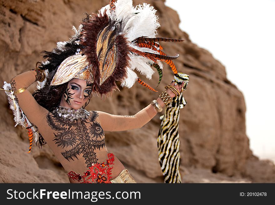 Tribal female wearing a feather head gear while holding a stripe cloth. Posing outside with a mountain ridge in the background