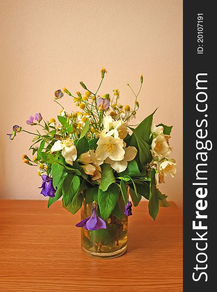 Seasonal wild flowers bouquet standing on a table in a glass with wall on a background. Seasonal wild flowers bouquet standing on a table in a glass with wall on a background