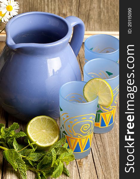 Composition with jug and three glass of lemonade, mint, half of lime and bunch of camomile on old wooden table. Composition with jug and three glass of lemonade, mint, half of lime and bunch of camomile on old wooden table