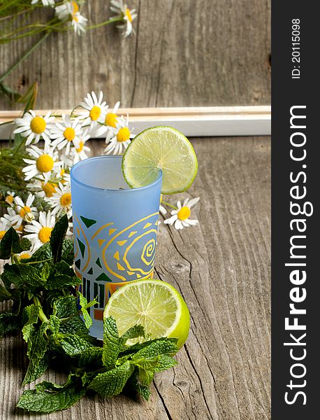 Composition with glass of lemonade, mint, half of lime and bunch of camomile on old wooden table. Composition with glass of lemonade, mint, half of lime and bunch of camomile on old wooden table