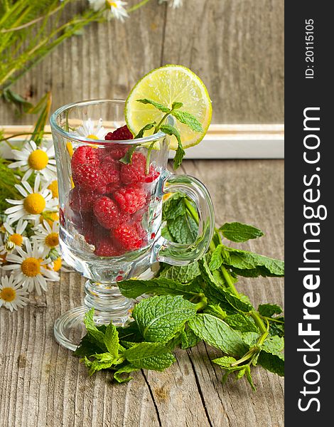 Still-life with fresh ripe raspberries in glass, bunch of camomile flowers and mint's leafes near mirror with reflection on old wooden table. Still-life with fresh ripe raspberries in glass, bunch of camomile flowers and mint's leafes near mirror with reflection on old wooden table