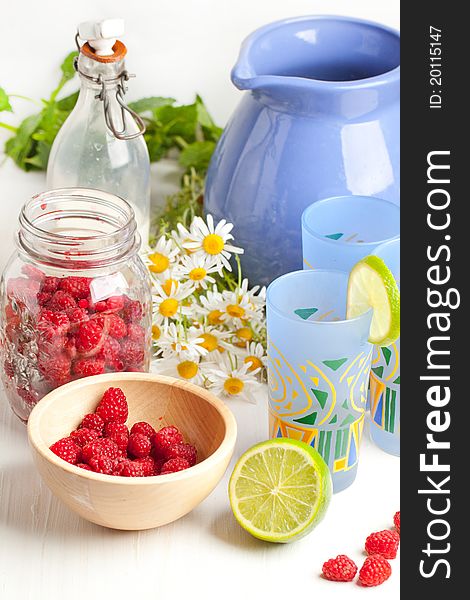 Composition with jug and three glasses of lemonade, fresh ripe raspberries, half of lime and bunch of camomile. Composition with jug and three glasses of lemonade, fresh ripe raspberries, half of lime and bunch of camomile