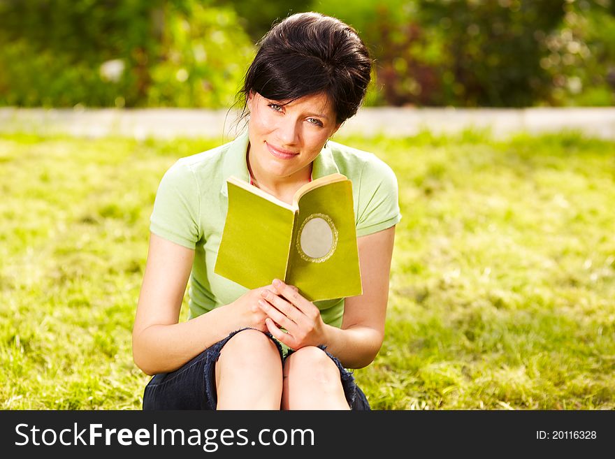 Caucasian woman is reading the green book sitting on the grass in the park. Caucasian woman is reading the green book sitting on the grass in the park