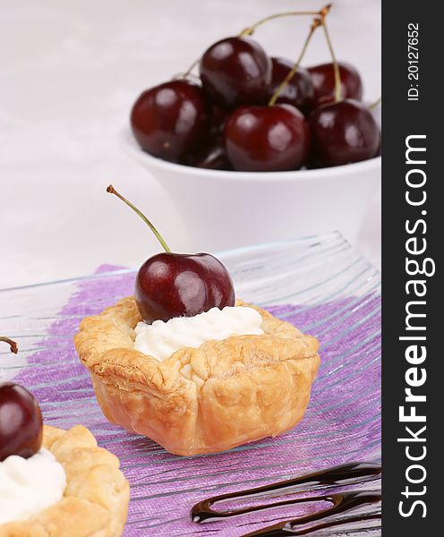 Small cherry tarts with whipped cream served on a glass plate. Cherryes out of focus in the background. Selective focus, shallow DOF.