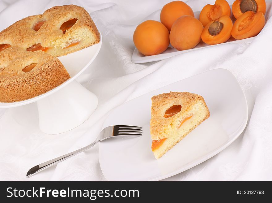 A slice of apricot tart served on a white plate. Apricot tart on a cake stand and apricots out of focus in the background