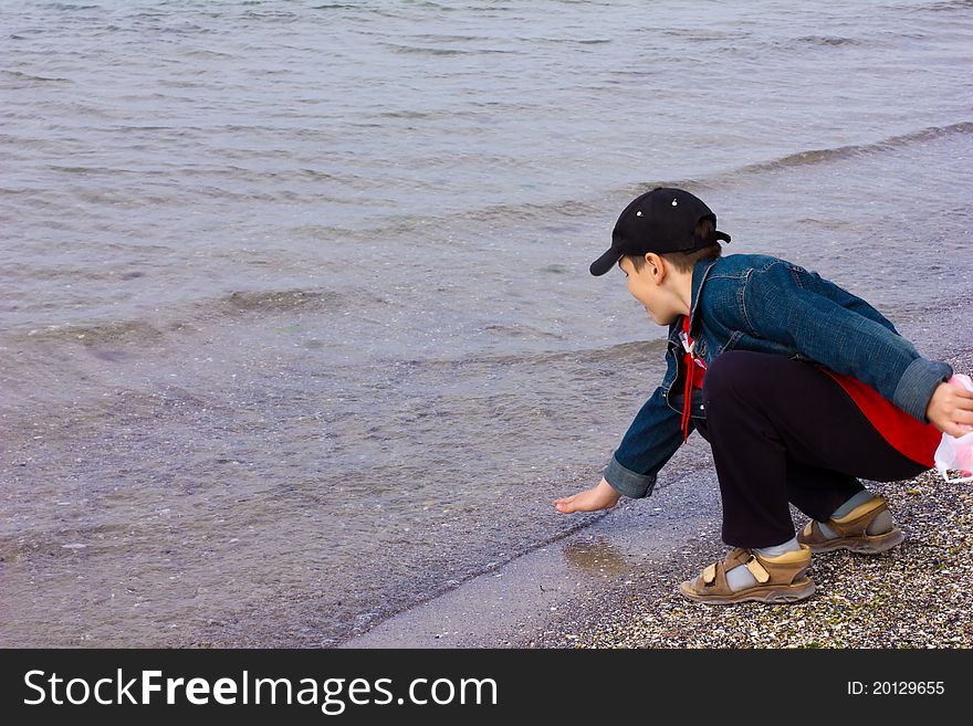 10 year old boy in denim jacket, cap, sandals squatting on the beach and the water touches his hand. 10 year old boy in denim jacket, cap, sandals squatting on the beach and the water touches his hand