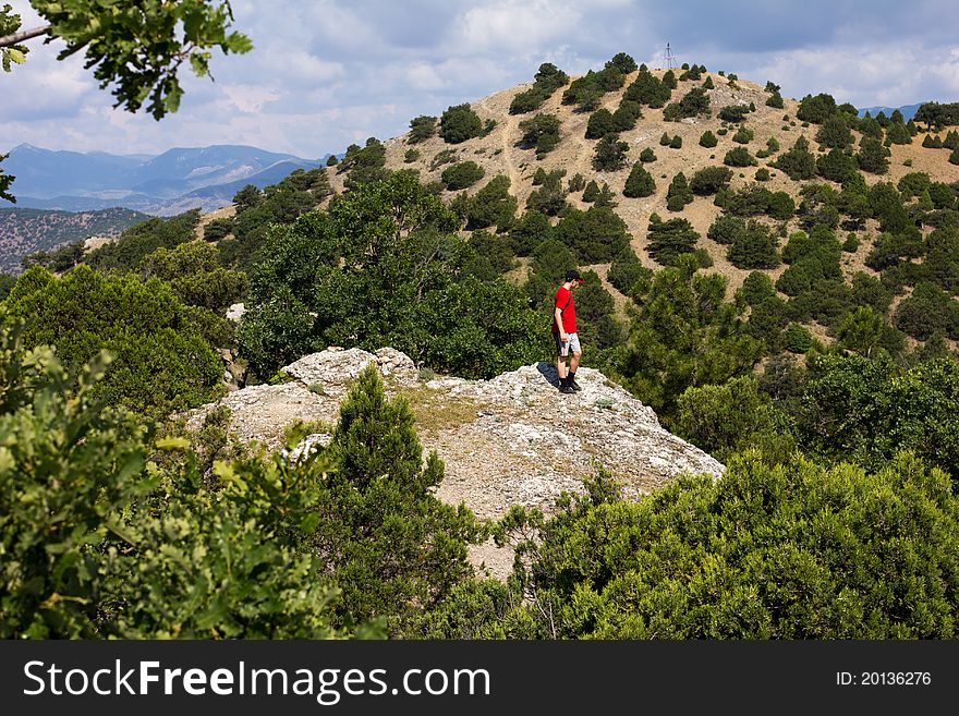 A teenager in a cap and a red shirt standing on the rocky top of a hill and looks to the next mountain. A teenager in a cap and a red shirt standing on the rocky top of a hill and looks to the next mountain