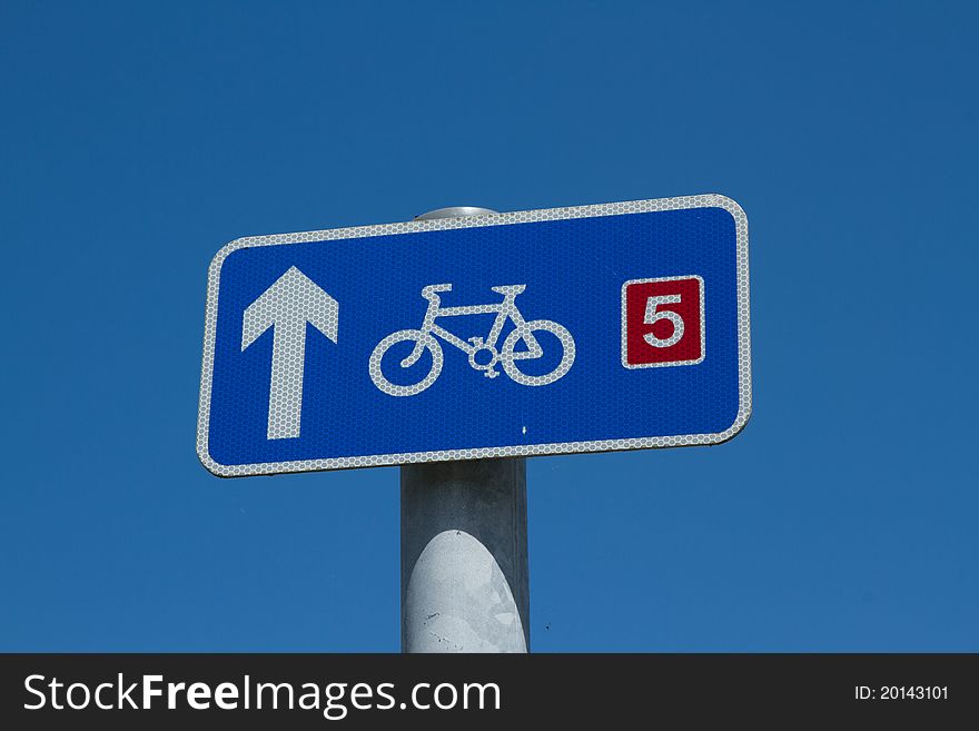 A blue sign with a white arrow and bicycle symbol and a route number five in red on a post against a blue sky. A blue sign with a white arrow and bicycle symbol and a route number five in red on a post against a blue sky.