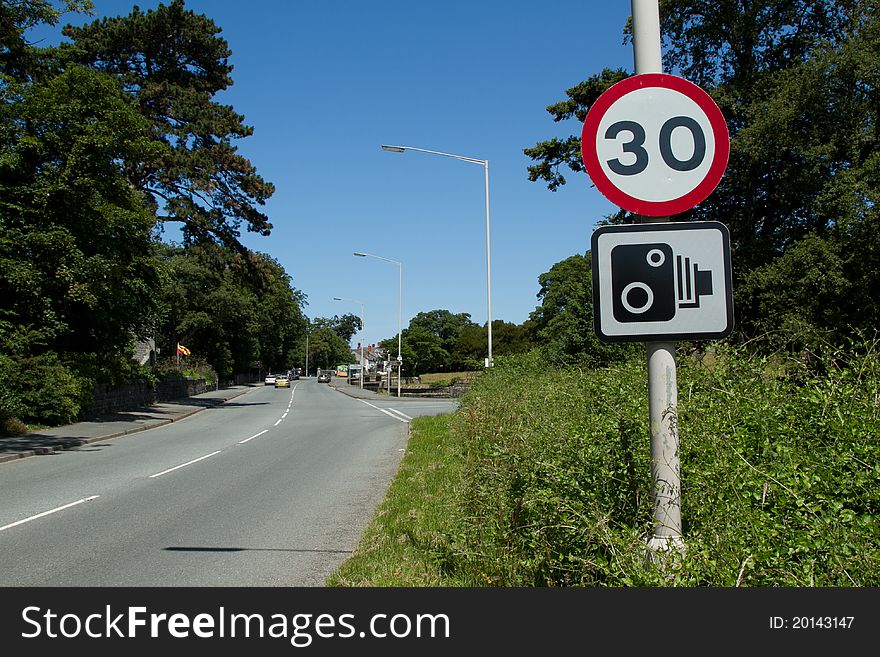 A thirty mile an hour sign and a speed camera sign on a post next to a road with cars in the distance. A thirty mile an hour sign and a speed camera sign on a post next to a road with cars in the distance.