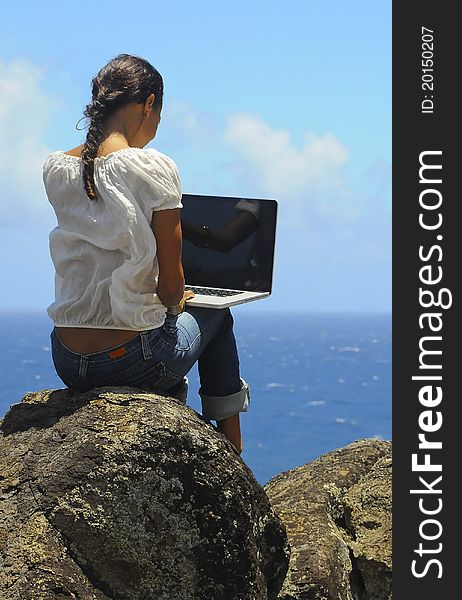 A young woman sits on a rock overlooking the ocean while working on her laptop computer. A young woman sits on a rock overlooking the ocean while working on her laptop computer