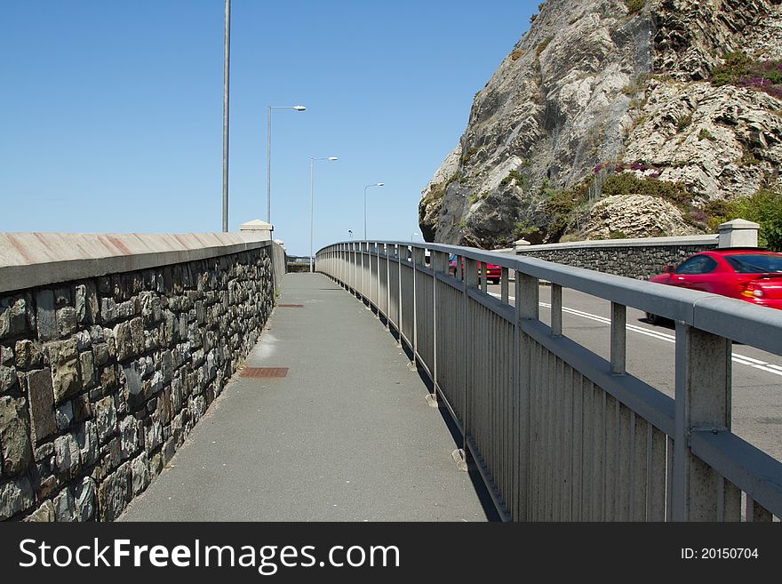 A pavement with a stone wall and lamp posts and metal railings separating the road with traffic. A pavement with a stone wall and lamp posts and metal railings separating the road with traffic.