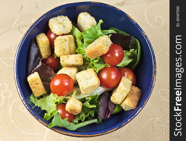 Summer Salad with summer greens, tomatos, croutons in a blue bowl with yellow cloth. Overhead view.