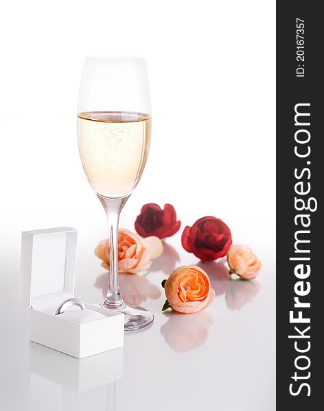 Champagne glass filled with Champagne surrounded by pink and red roses and a white jewelry box with a ring in it on a white background. Champagne glass filled with Champagne surrounded by pink and red roses and a white jewelry box with a ring in it on a white background