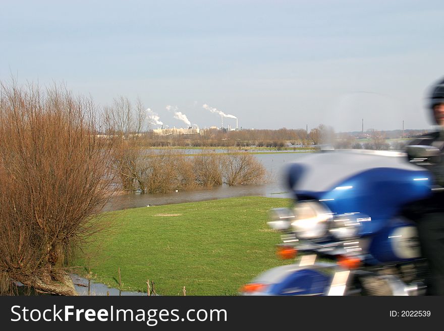 A motorcyclist and smoking factory-chimneys in a dutch riverscape. The forelands are swamped. A motorcyclist and smoking factory-chimneys in a dutch riverscape. The forelands are swamped.