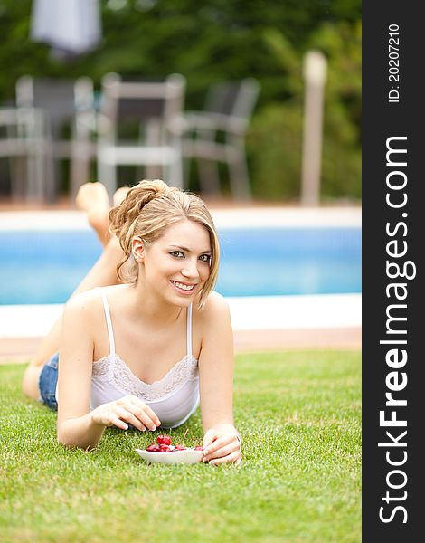 Young beautiful blonde woman eating cherries on the grass smiling. Young beautiful blonde woman eating cherries on the grass smiling