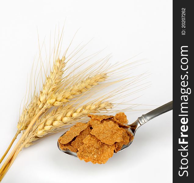 Wheat heads next to a spoonful of wheat cereal flakes on a white background