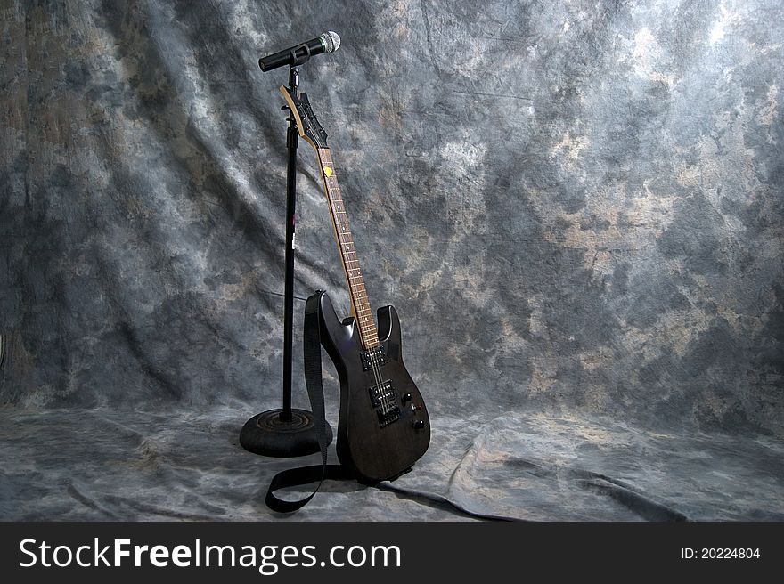 A black electric guitar is resting on a microphone stand with a microphone attached. A black electric guitar is resting on a microphone stand with a microphone attached.