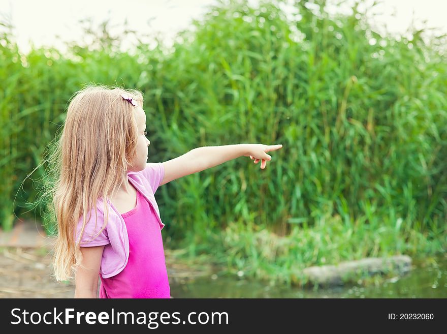 Blonde little girl shows forefingers aside near river with canes in summer