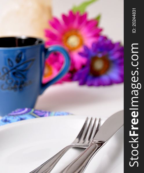 Coffee up with white plate, knife, and fork. Blurred purple and pink flowers in the background. Coffee up with white plate, knife, and fork. Blurred purple and pink flowers in the background.